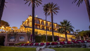 Een groot, meerijlaags hotel met een goed verlichte gevel, palmbomen en een aangelegde tuin met bloemen.