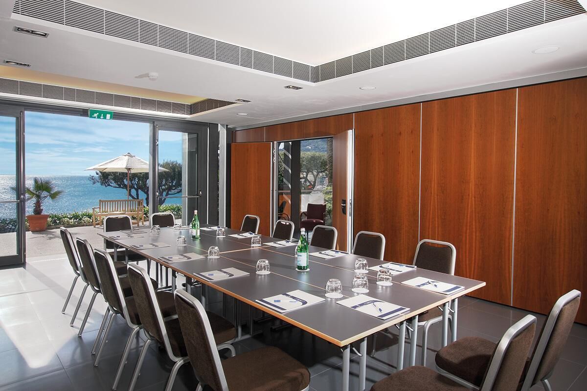 A conference room with a long table, chairs, and a view of the sea through large windows.