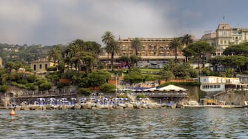 A coastal hotel with a view of the sea, palm trees, and a large building in the background.