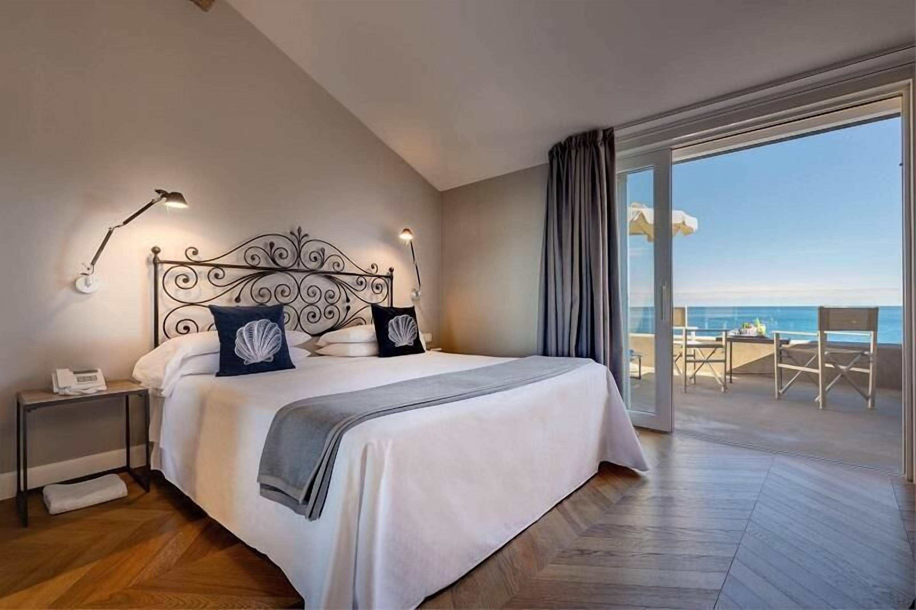 Una habitación de hotel moderna con una cama grande, una mesita de noche con lámpara y vista al océano a través de una puerta corrediza de vidrio.