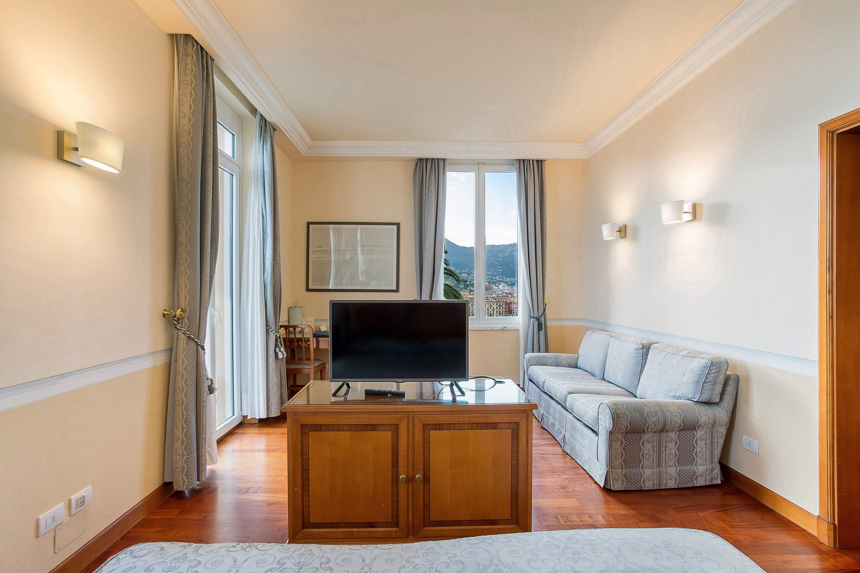 Ein Zimmer mit Flachbildfernseher auf einer Glastisch, einer Couch, einem Holzkabinett und Blick ins Freie durch ein Fenster.