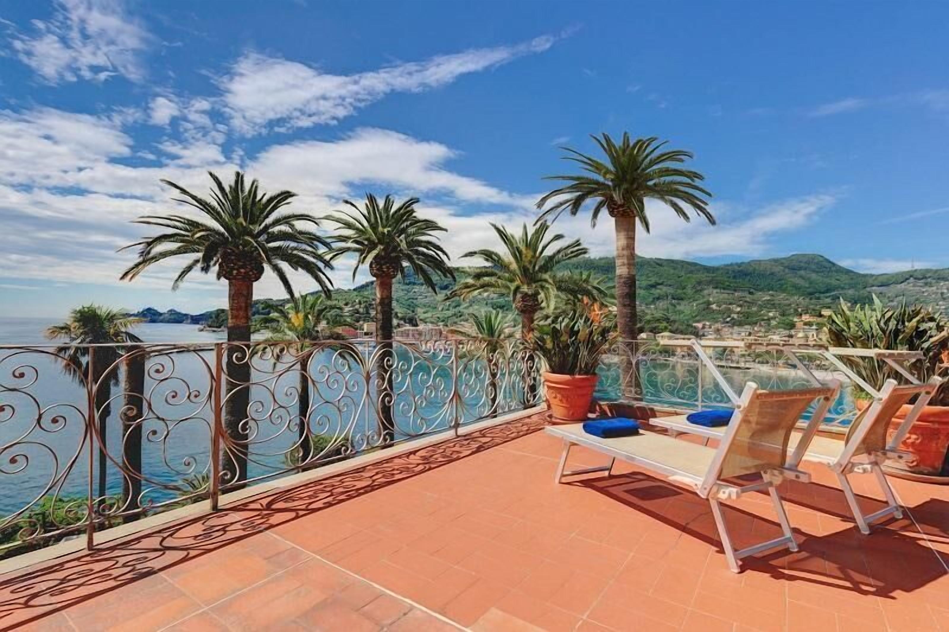 A terrace with a view of the sea, palm trees, and a pool area with lounge chairs.