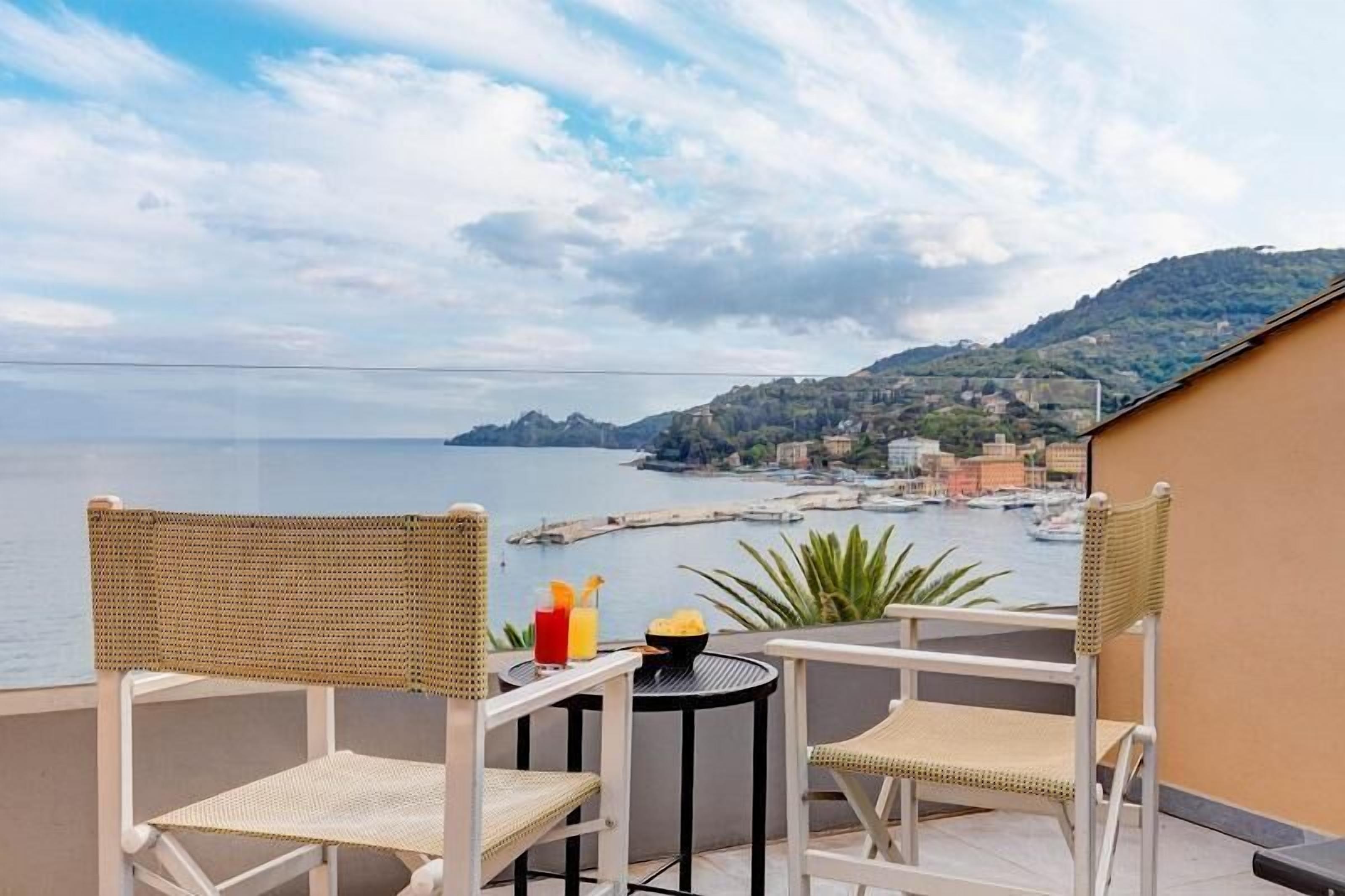Ein Balkon mit Korbstühlen, einem kleinen runden Tisch mit Getränken und Blick auf das Meer und eine Stadt.
