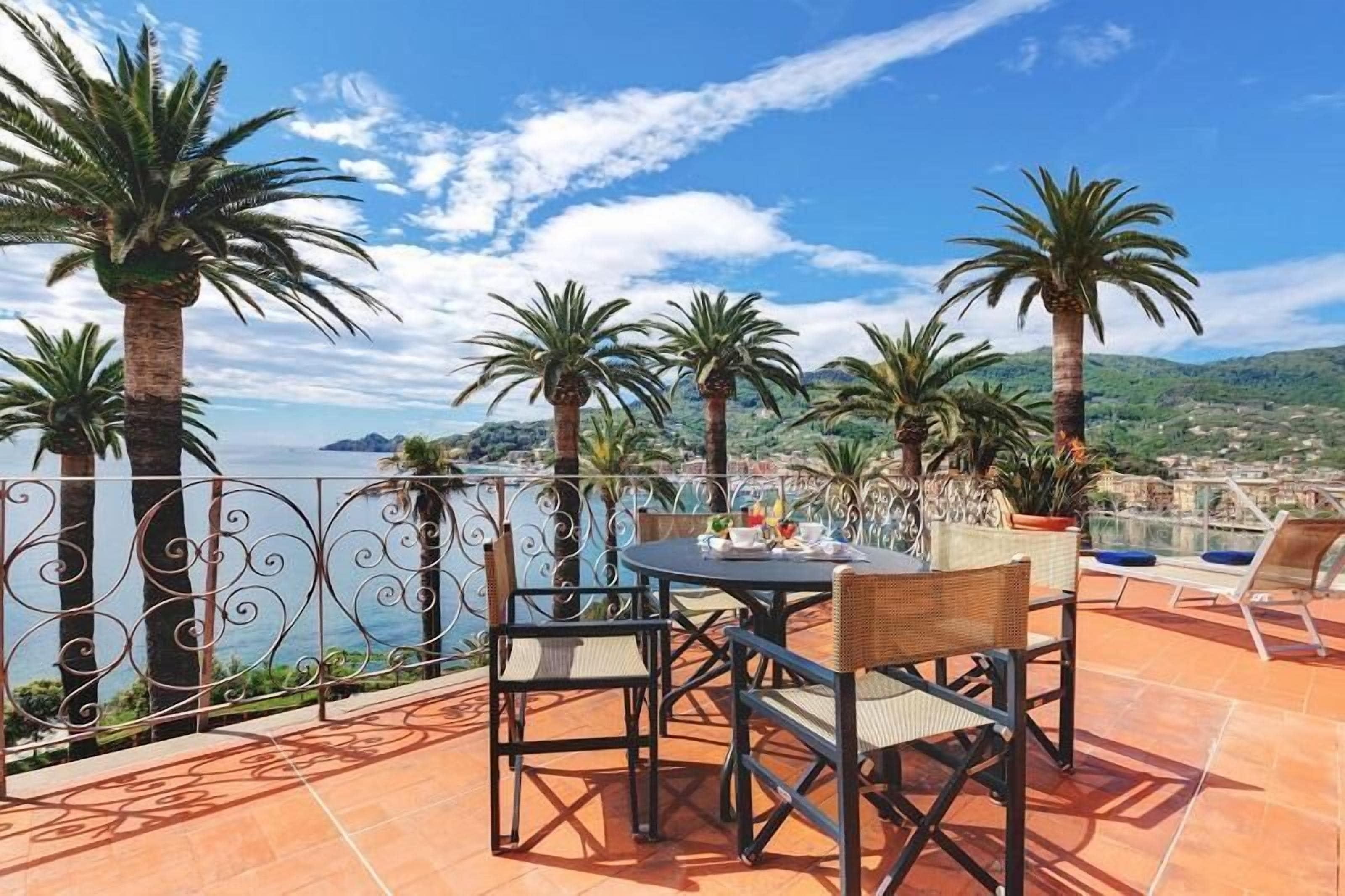 Terraço ao ar livre com mesa e cadeiras, rodeado por palmeiras e vista para o mar.