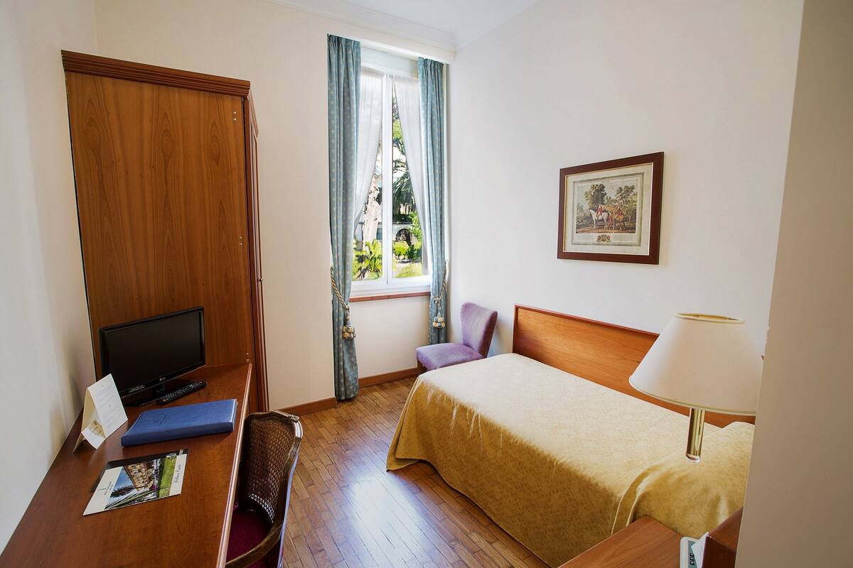Een hotelkamer met een houten bureau, een bed met een beige deken, een stoel, een televisie en een raam met gordijnen.