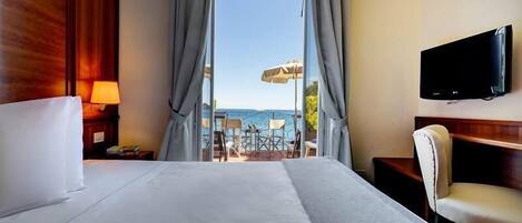 A hotel room with a large bed, a TV mounted on the wall, and a view of the beach through the window.