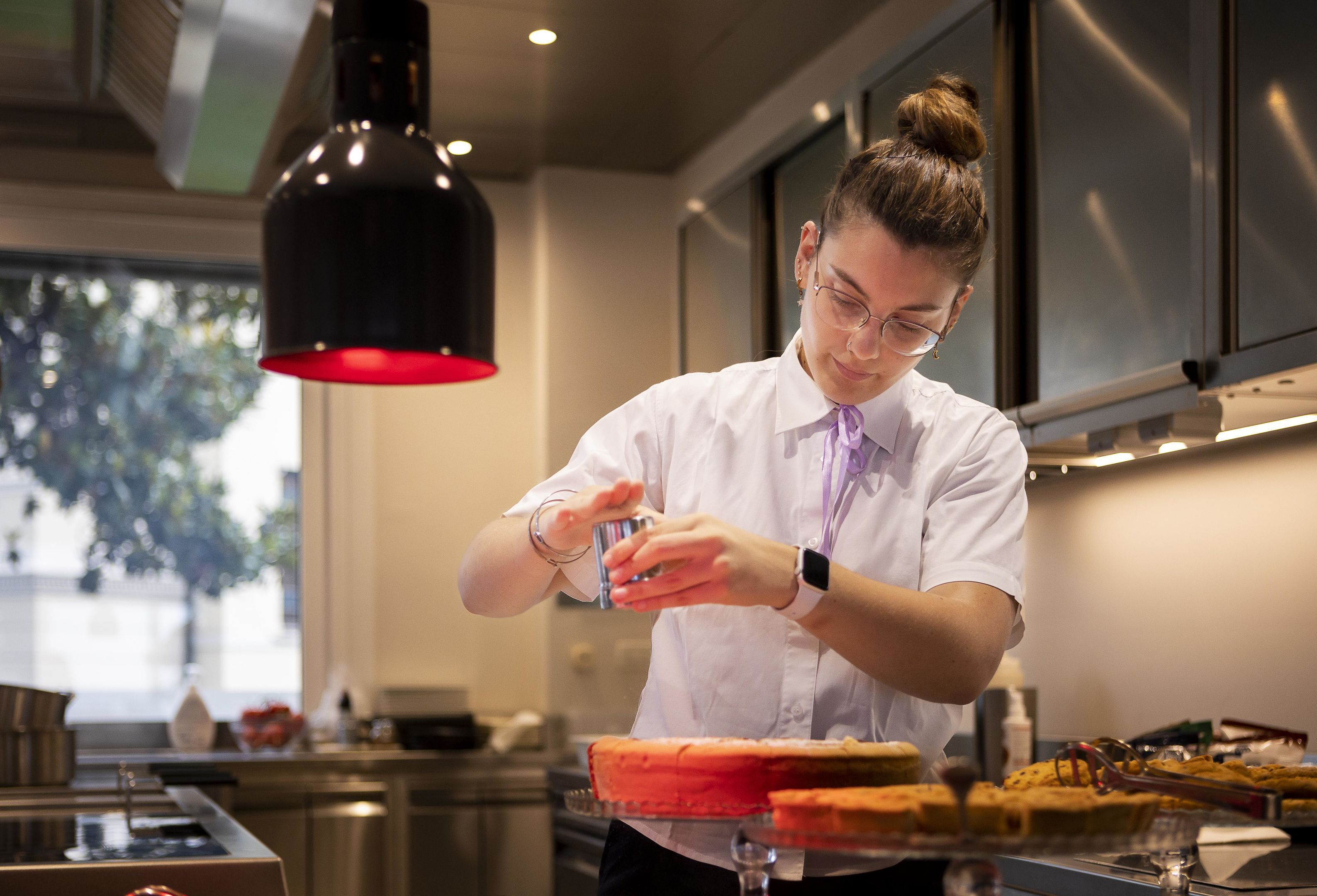 Una persona con camisa blanca y corbata morada preparando comida en una cocina moderna con electrodomésticos de acero inoxidable y una lámpara colgante roja.
