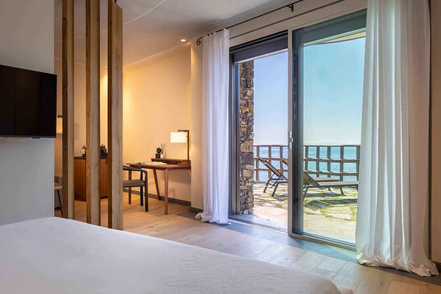 Una camera d'albergo con un letto, una scrivania e vista sul mare attraverso una porta-finestra in vetro.