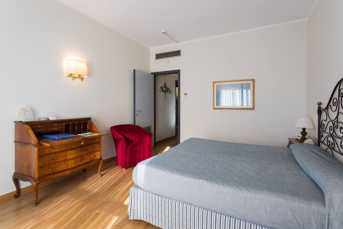 Een slaapkamer met een bed, een houten dressoir, een rode stoel, een lamp op de muur en een deur.