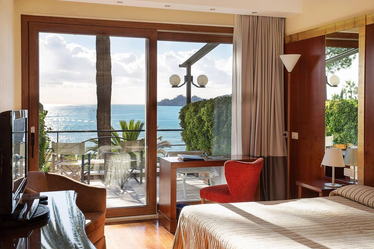Hotelkamer met een groot schuifglasraam dat leidt naar een balkon met uitzicht op de zee. Er is een bed, een bureau en een rode stoel.