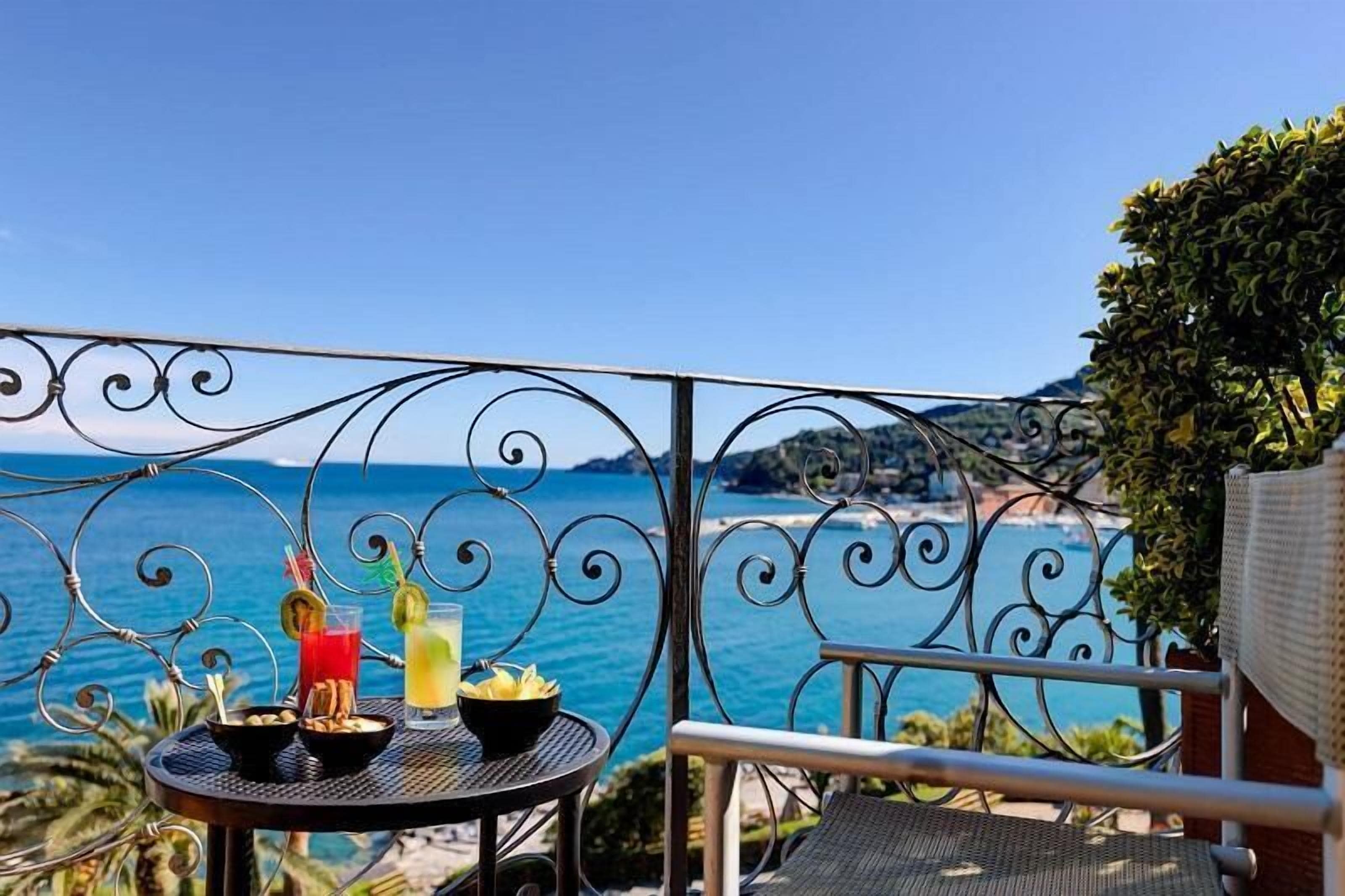 Un balcón con barandilla metálica, una mesa redonda con bebidas y aperitivos, y vistas al mar y a las montañas.