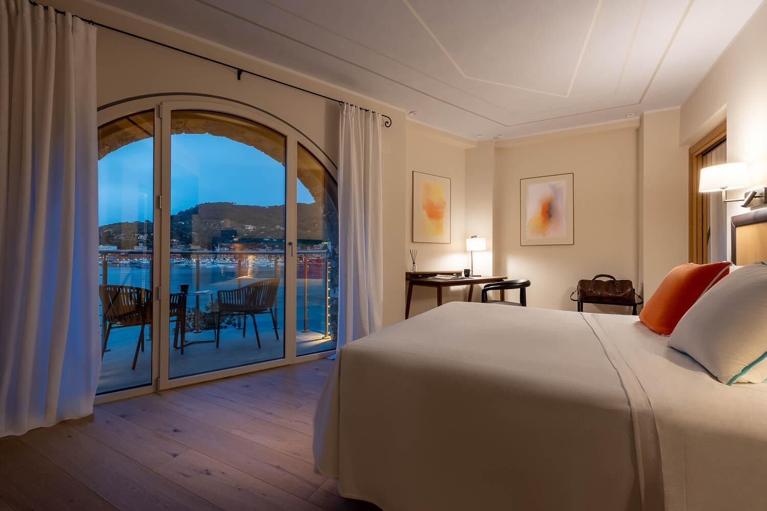 A hotel room with a large bed, a desk, a chair, and a balcony with a view of the city.