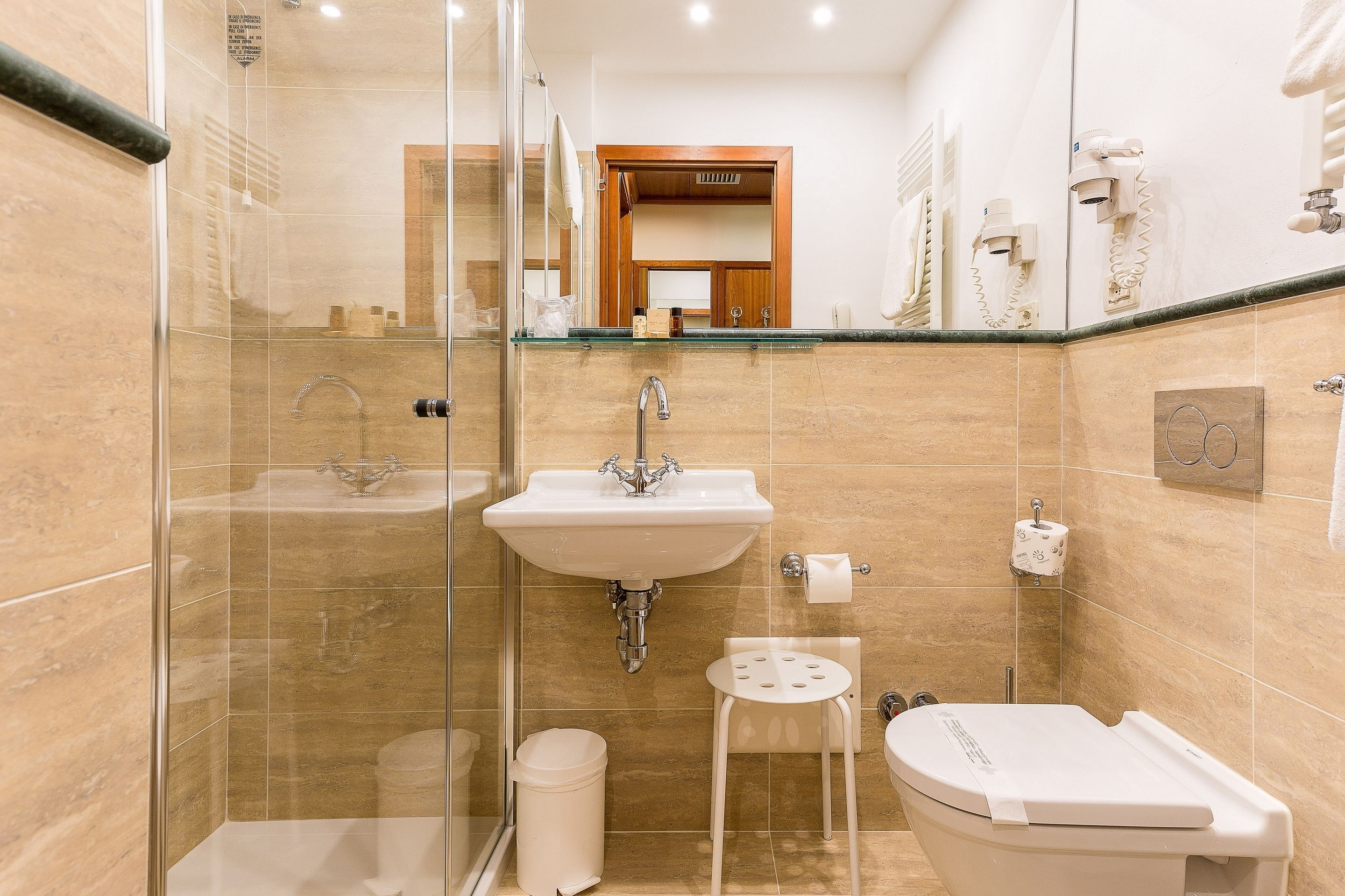 A bathroom with a glass-enclosed shower, a sink, a toilet, and a small stool.