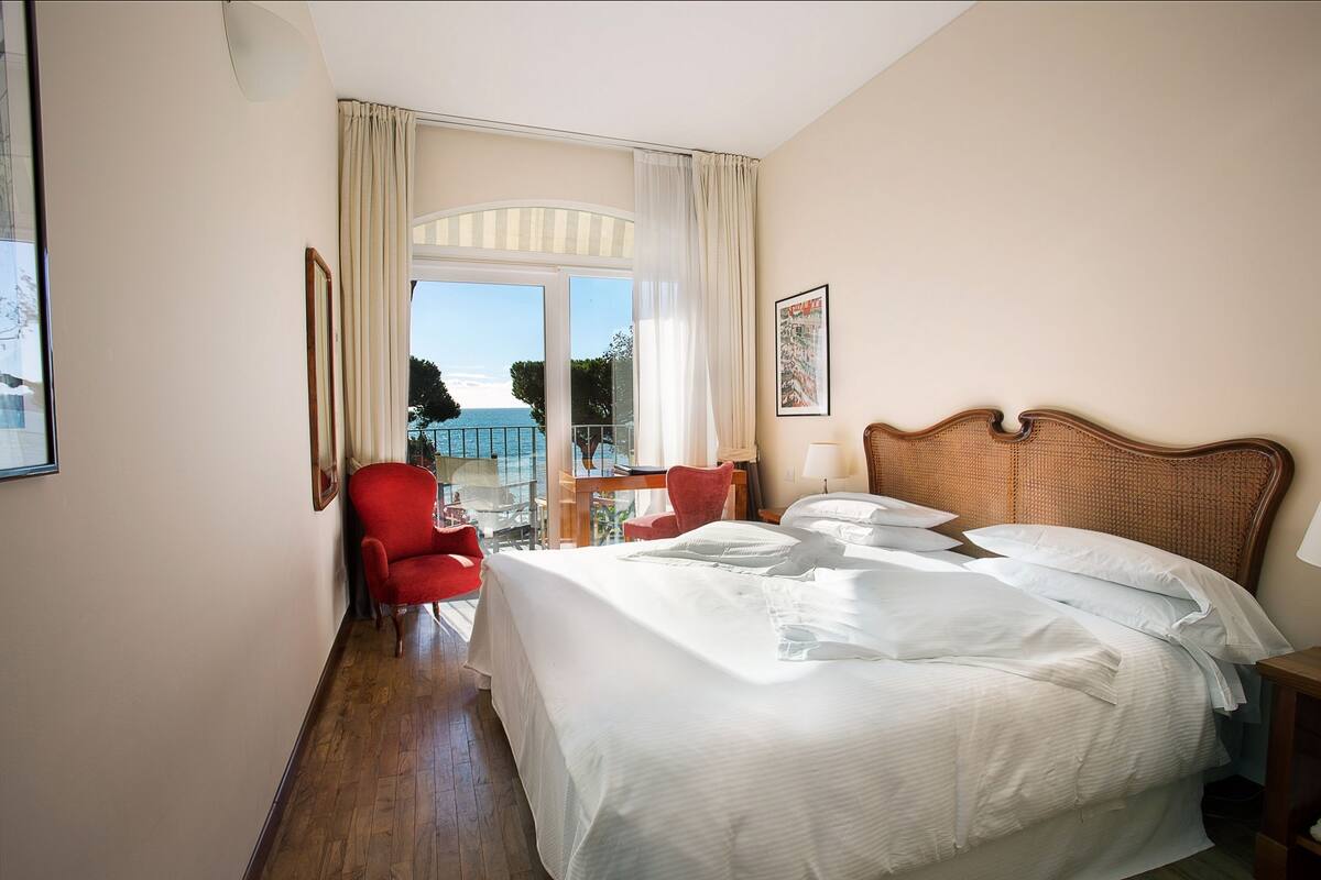 Hotelkamer met een groot bed, een rode stoel, een nachtkastje van hout en uitzicht op zee door een raam met gordijnen.