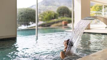 A person in a hot tub with a waterfall feature, surrounded by a pool and a view of greenery and mountains.