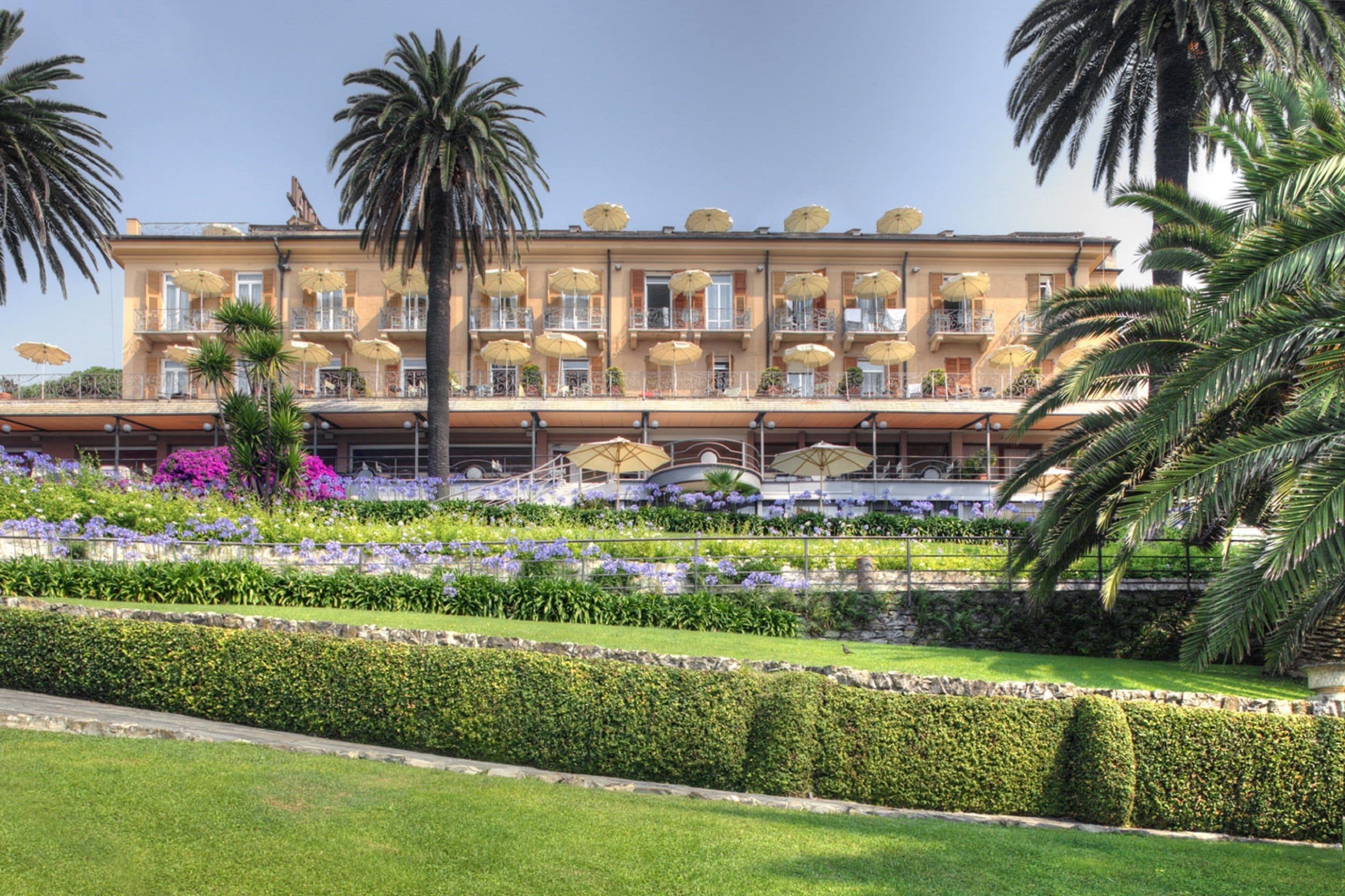 A hotel with a symmetrical facade, multiple balconies, and a well-maintained garden with palm trees and flowers.