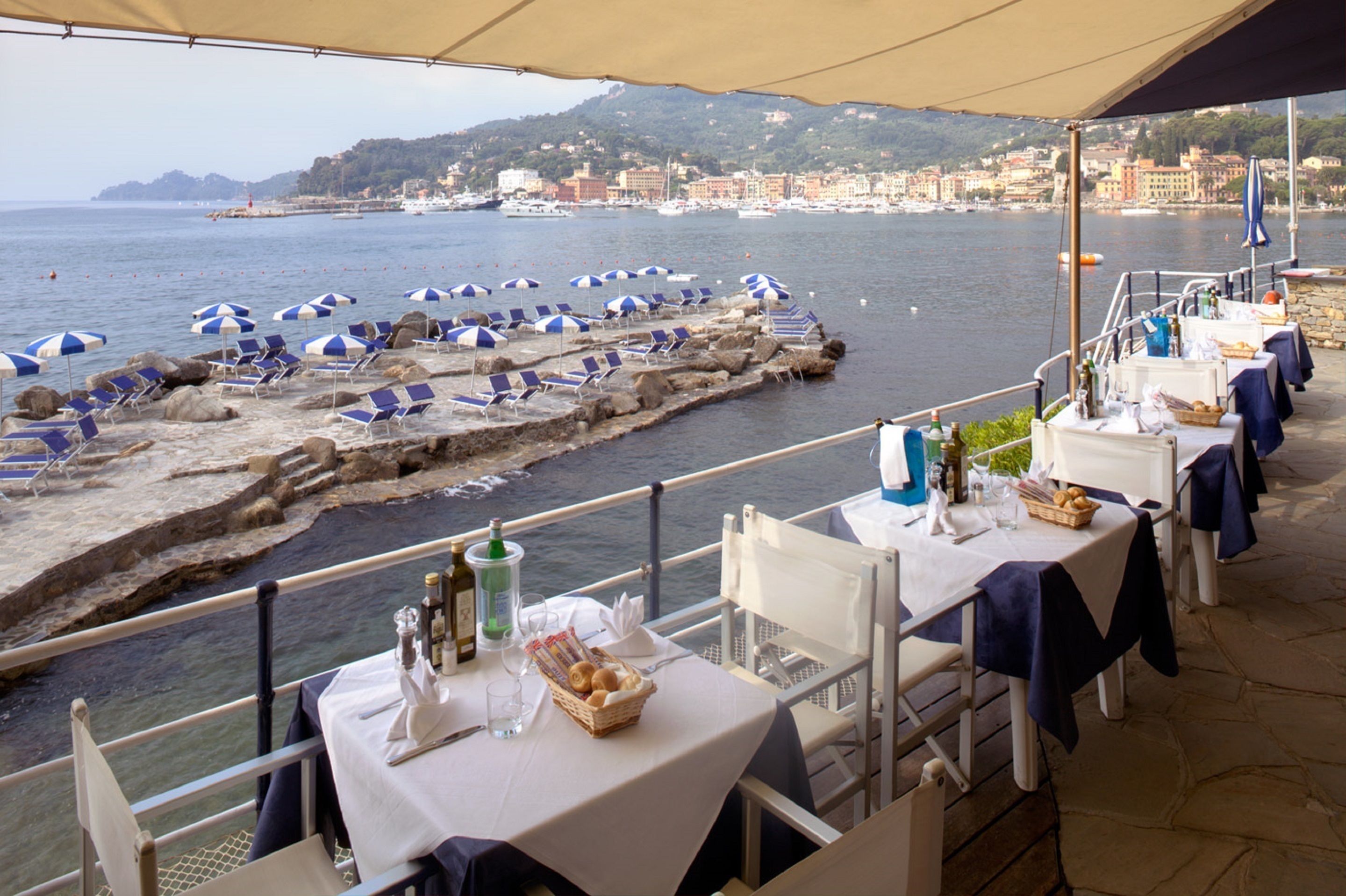 A terrace with tables and chairs overlooking a rocky waterfront with beach umbrellas.