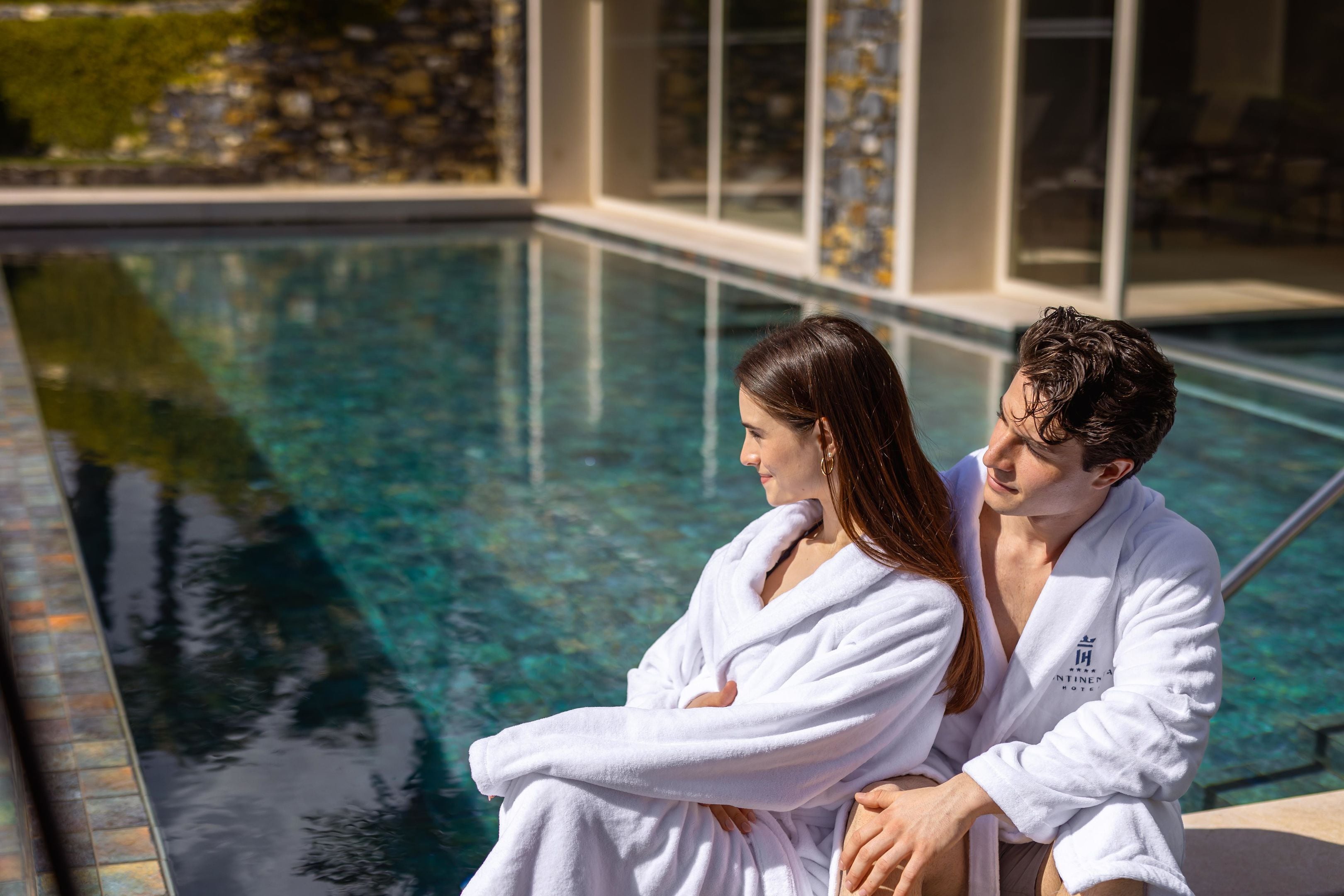 Un couple est assis au bord d’une piscine dont l’eau est cristalline, entouré d’un bâtiment moderne doté de grandes fenêtres et d’une façade en pierre.