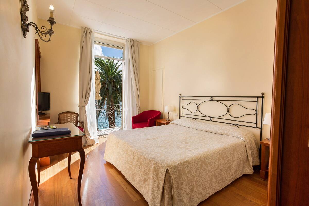 Een slaapkamer met een bed, een bureau, een stoel, een televisie en een balkon met uitzicht op palmbomen.