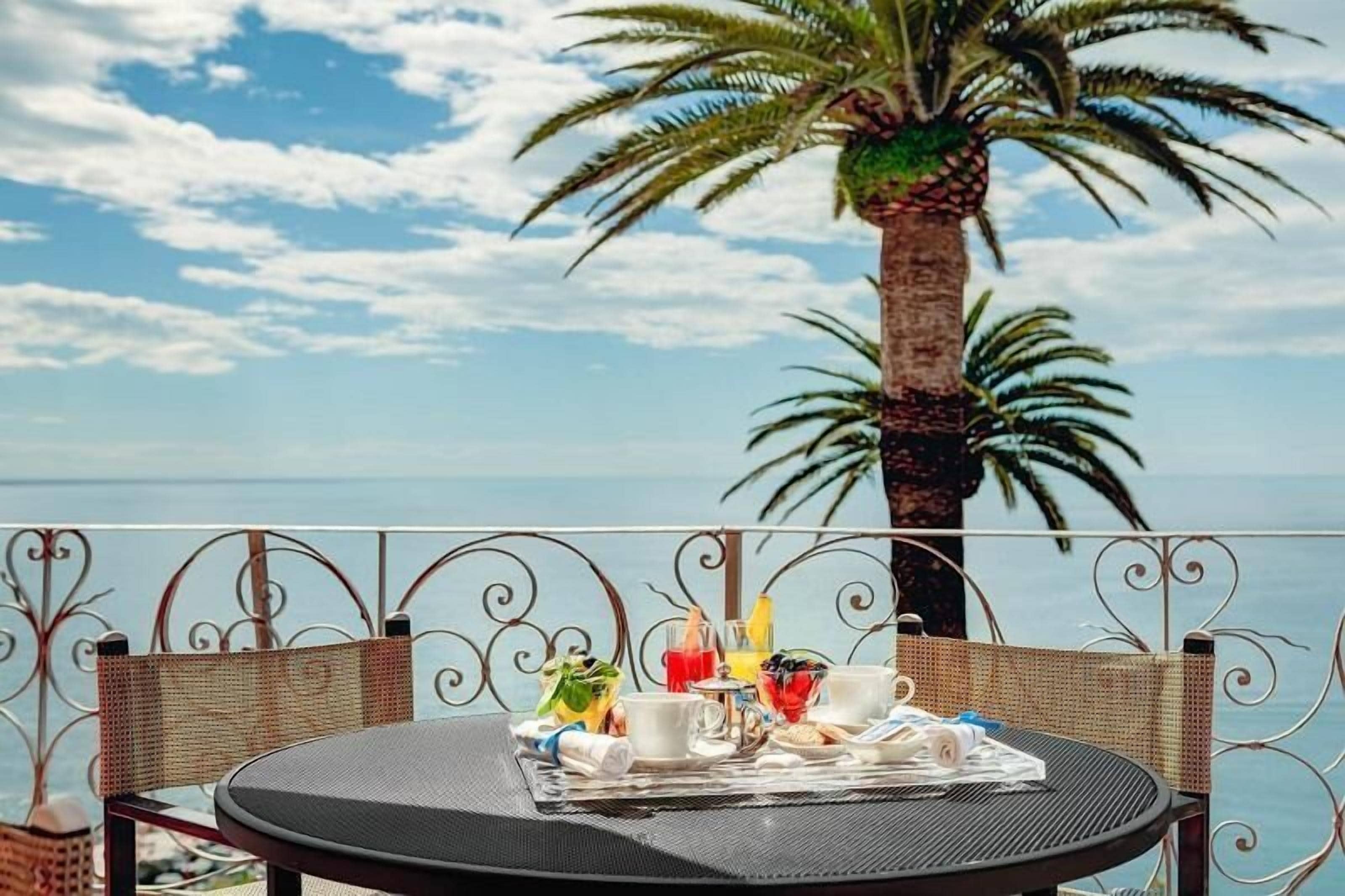A round table with a metal frame, set with a newspaper, cups, and a glass of juice, overlooking a palm tree and the sea.