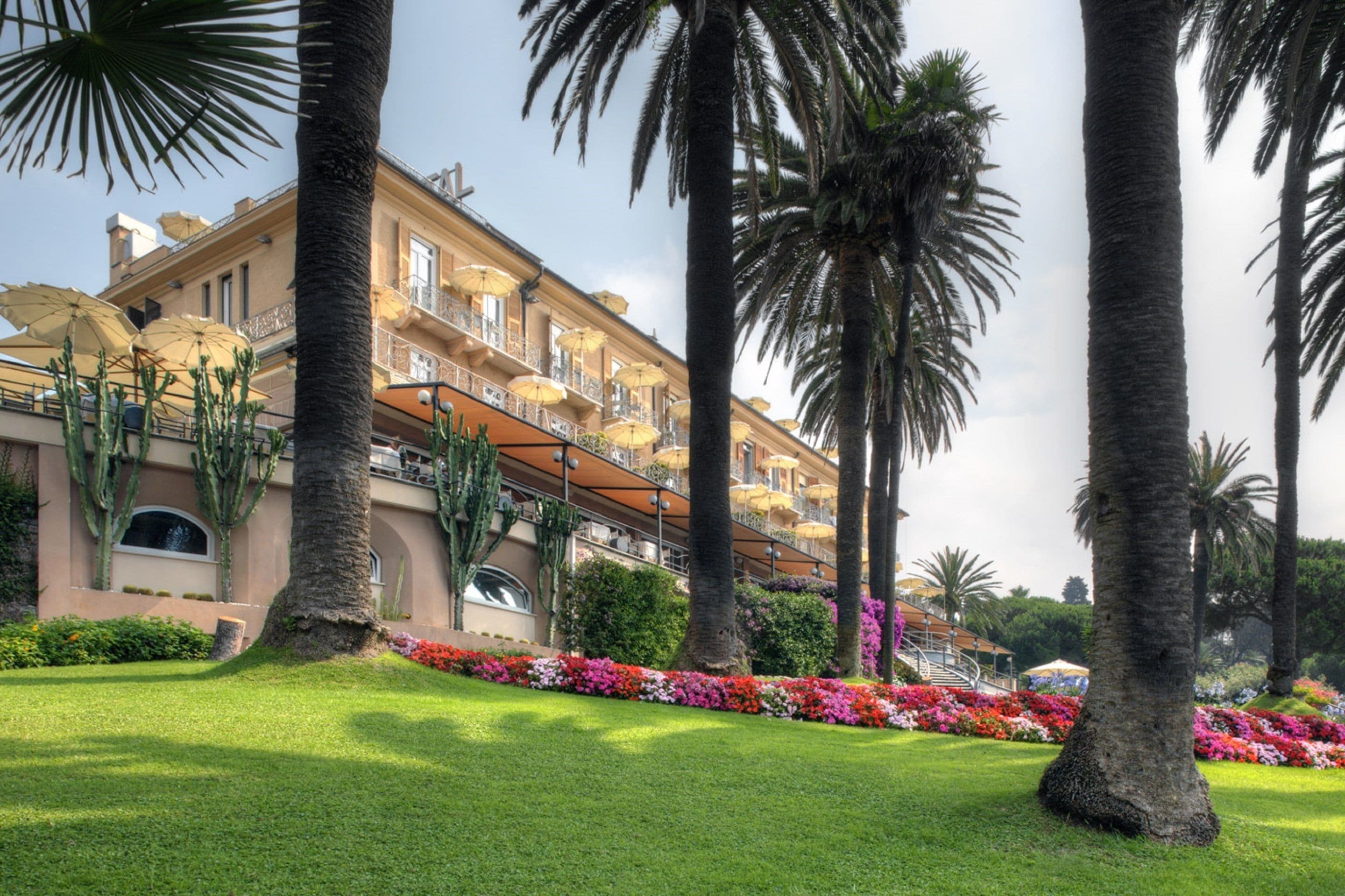 A hotel with a well-maintained garden, palm trees, and a balcony with umbrellas.