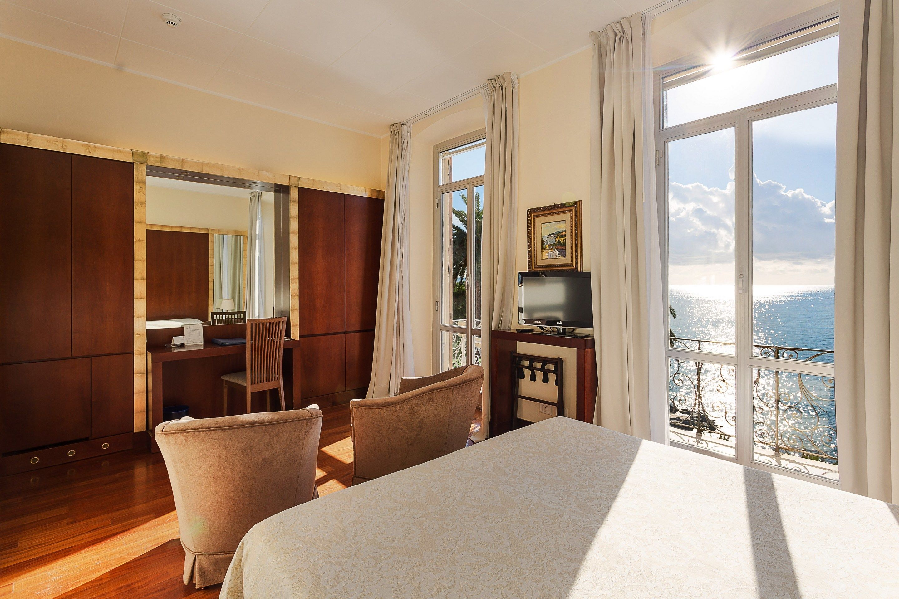 A hotel room with a large bed, a desk with a chair, a television, and a view of the sea.