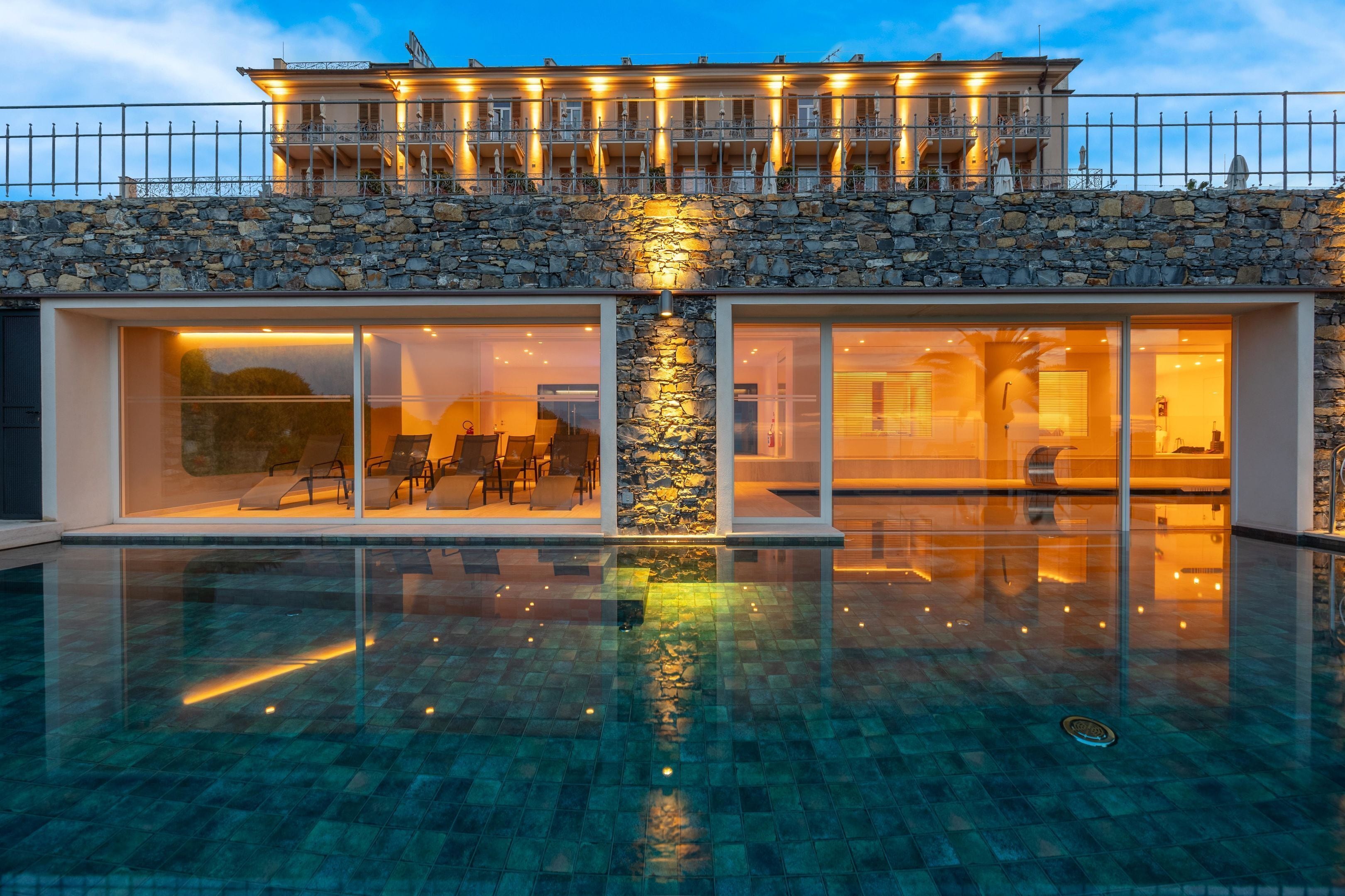 A modern building with a stone facade, large glass windows, and a swimming pool in front.