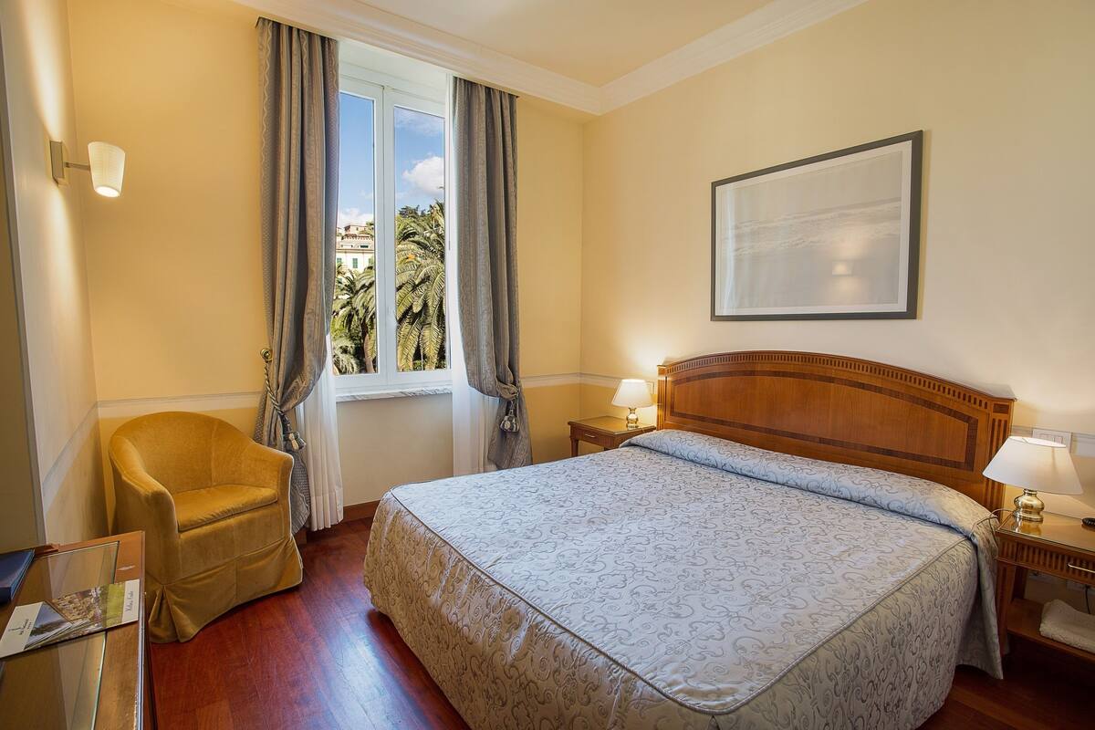 Een hotelkamer met een groot bed, een houten hoofdbord, een stoel, een raam met gordijnen en een ingelijst schilderij aan de muur.