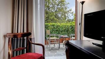A hotel room with a wooden chair, a red cushion, a black TV, and a view of a garden through a window.