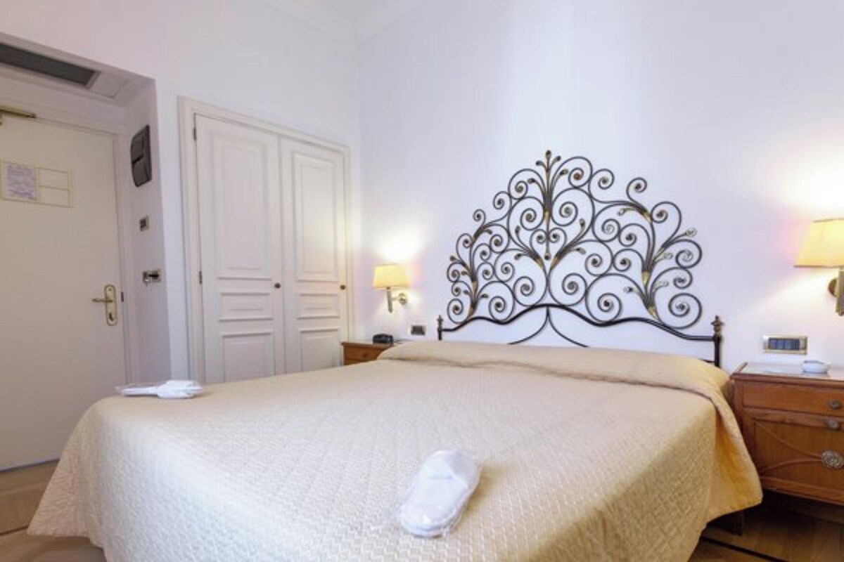 Ein ordentlich bezogenes Bett mit dekorativem Kopfteil, ein Nachttisch mit Lampe und eine Tür mit Schloss.