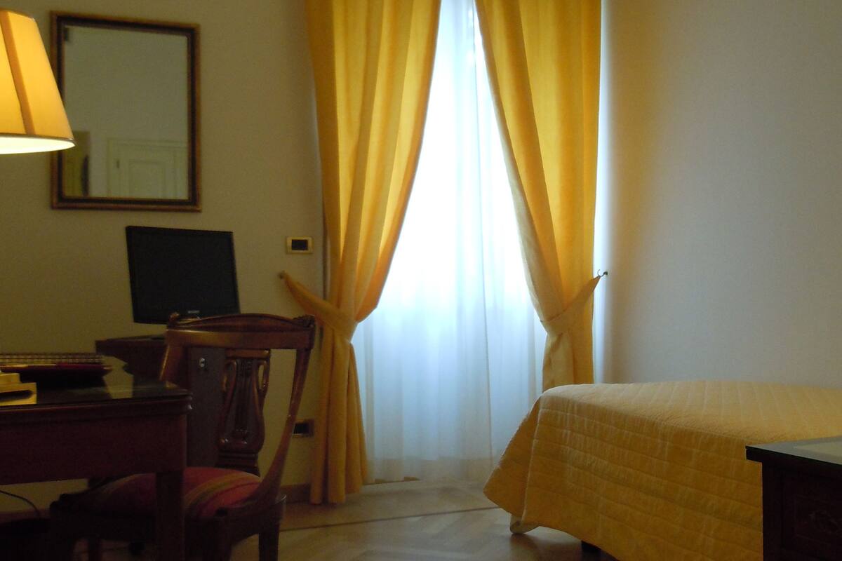 Une chambre d’hôtel avec un lit, un bureau doté d’une lampe, une chaise, un ordinateur et une fenêtre avec des rideaux jaunes.