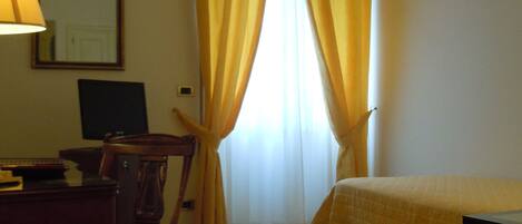 A hotel room with a bed, a desk with a lamp, a chair, a computer, and a window with yellow curtains.