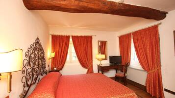 A bedroom with a large bed, wooden beams, a desk with a chair, and two windows with red curtains.