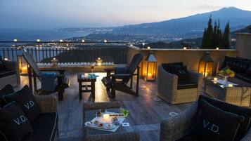 A rooftop terrace with wicker furniture, candles, and a view of a city and mountain at dusk.