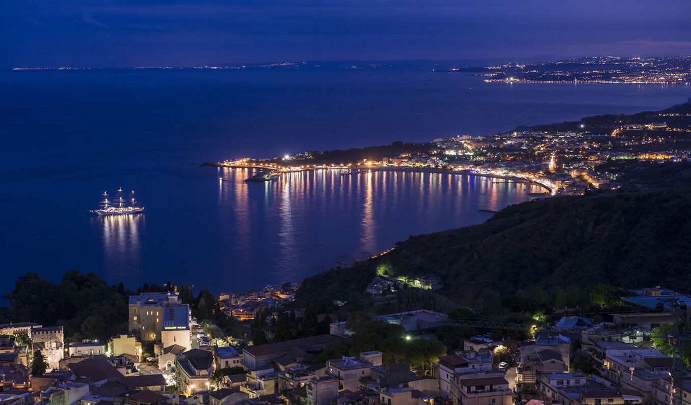 Un pueblo costero de noche con un paseo marítimo destacado, un puente y una estructura similar a un castillo.