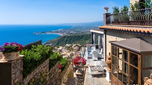 A coastal building with a stone wall, a balcony with a railing, and a view of the sea and distant town. - Hotel Villa Ducale (Taormina)