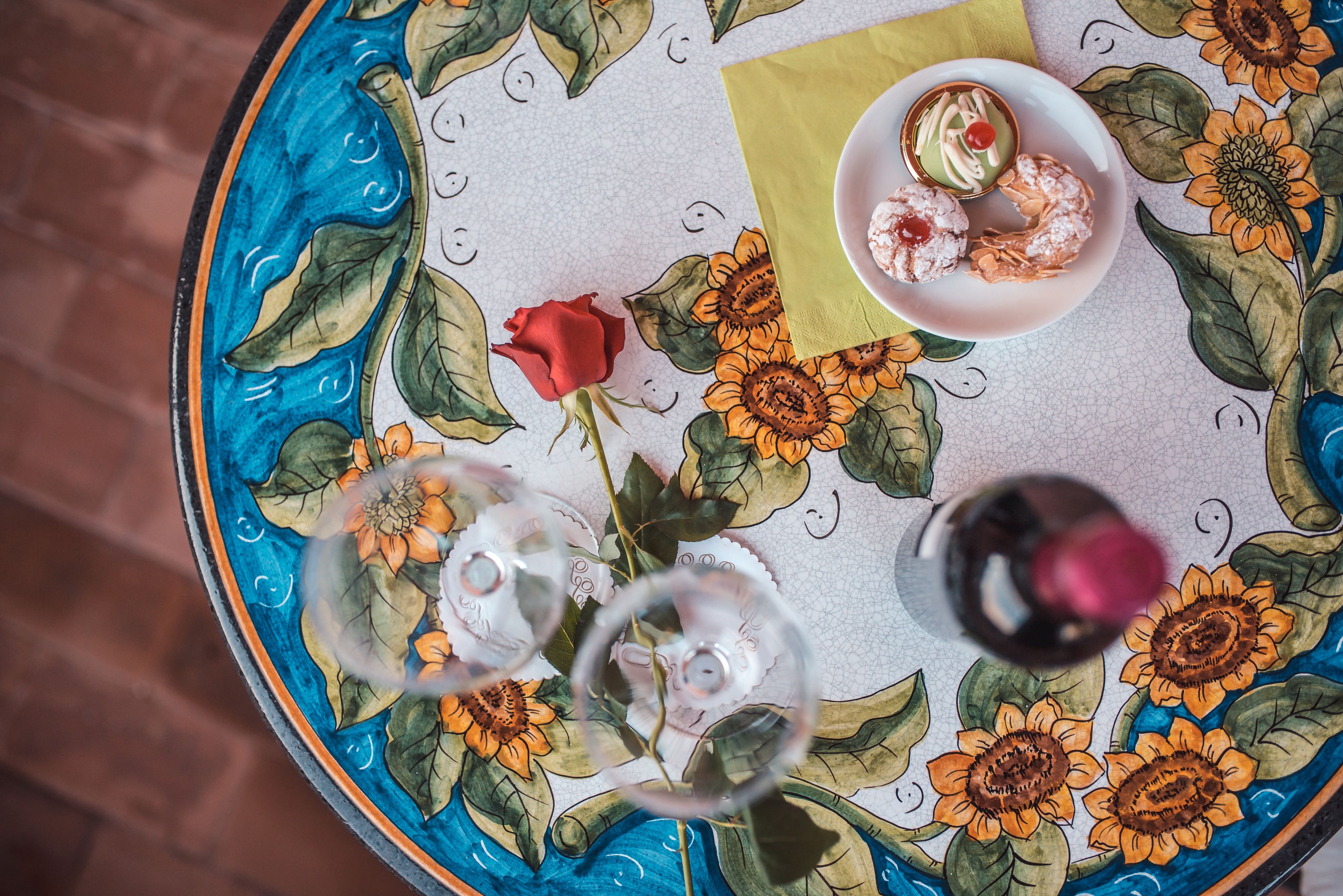 Une table ronde recouverte d’une nappe à motifs floraux, une rose et une assiette remplie de pâtisseries, ainsi qu’un verre de vin rouge.