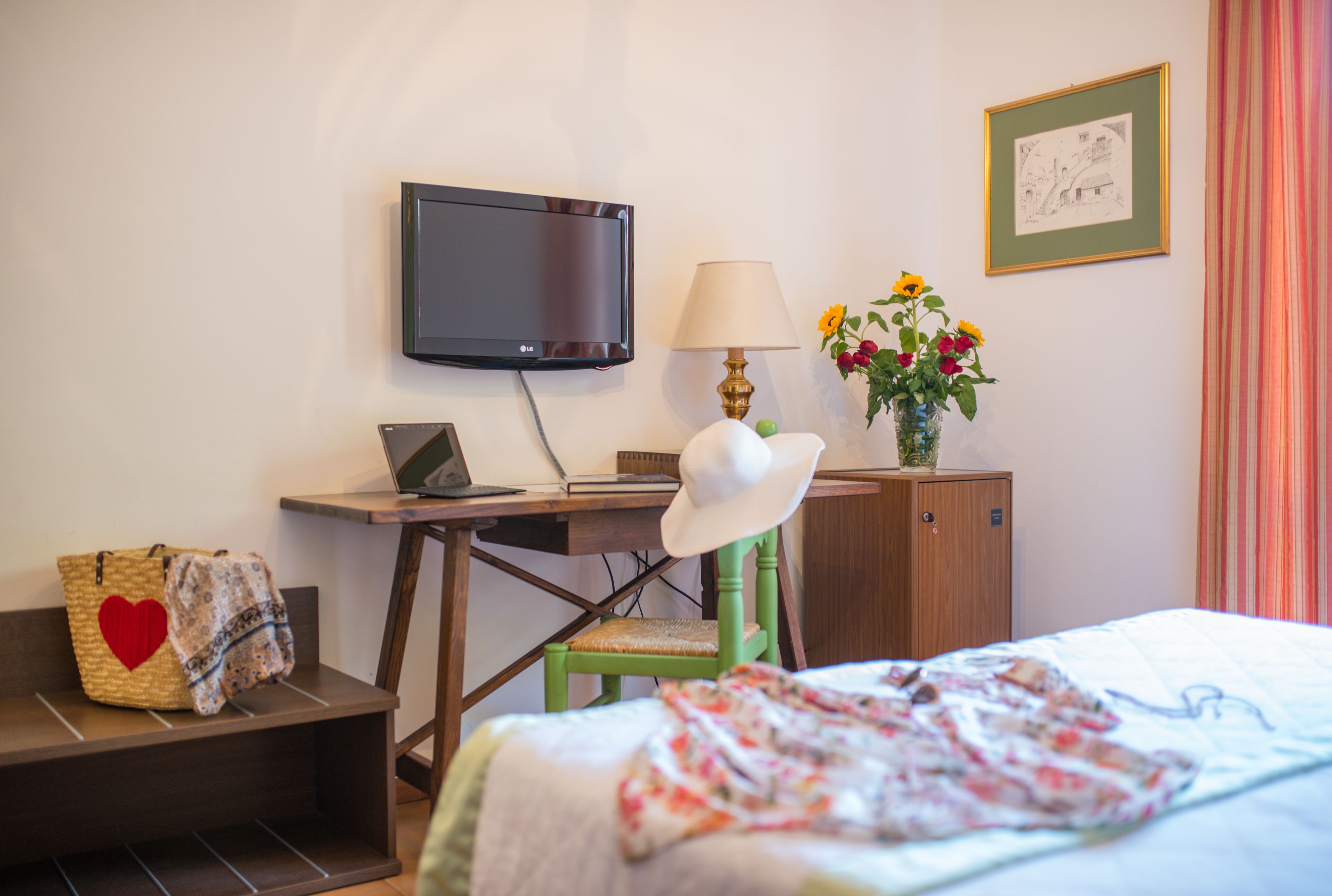 Ein Hotelzimmer mit Bett, Schreibtisch mit Laptop, Fernseher, Lampe, Stuhl, Schrank und einer Blumenvase.