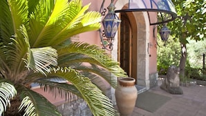 Una palmera en primer plano, una maceta de piedra y un edificio con una cúpula azul y dos faroles.