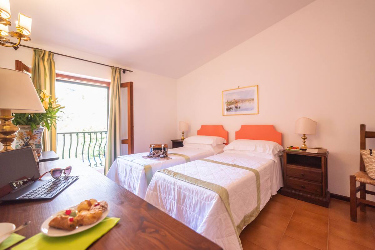 Une chambre d’hôtel avec deux lits, un bureau avec un ordinateur portable et des viennoiseries, une chaise et un balcon avec des rideaux.