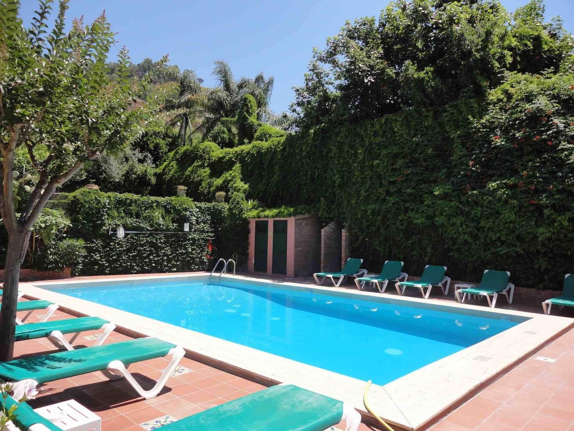 Una piscina rectangular con borde de azulejos, rodeada de sillas reclinables verdes y un muro de ladrillo.