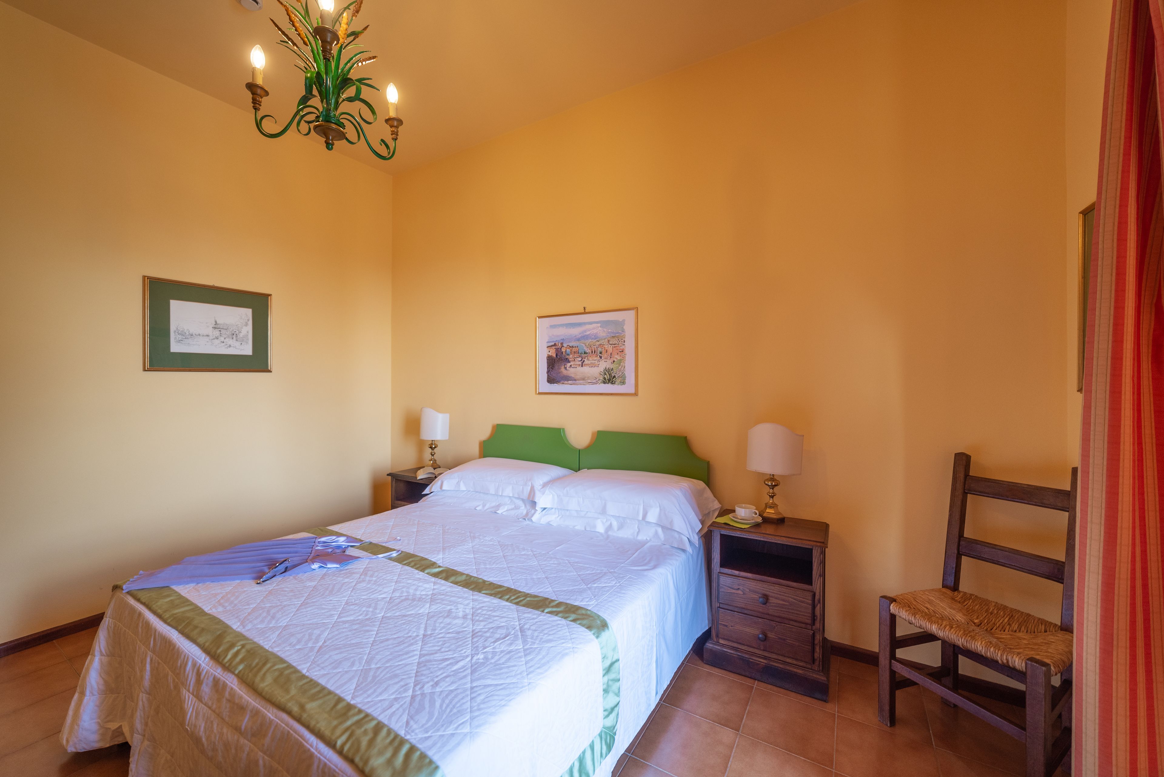 Ein Hotelzimmer mit einem Bett, zwei Nachttischen, einem Sessel, einer Deckenleuchte und zwei gerahmten Bildern an den Wänden.
