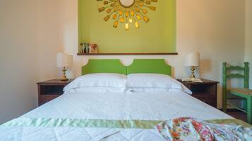 Un lit bien fait, avec du linge de lit blanc, deux tables de chevet avec des lampes, une tête de lit verte et un miroir mural orné d’un motif en forme de soleil.