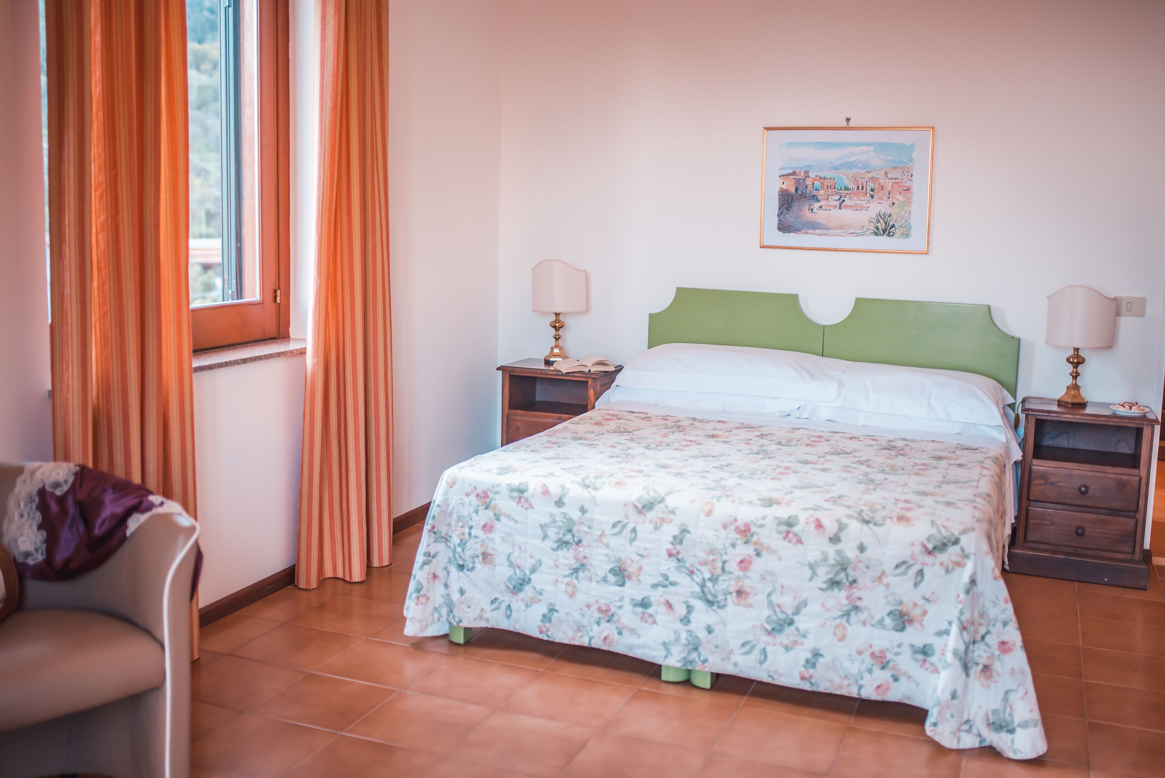Un dormitorio con cama, dos mesitas de noche, una silla, cortinas, ventana y un cuadro colgado en la pared.