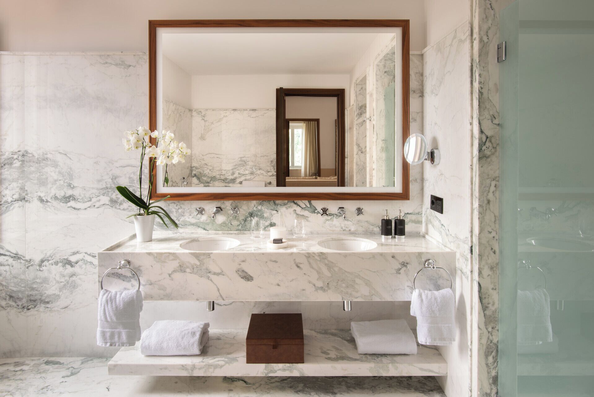 Bagno con due lavabi, uno specchio grande, pareti in marmo e cabina doccia in vetro.