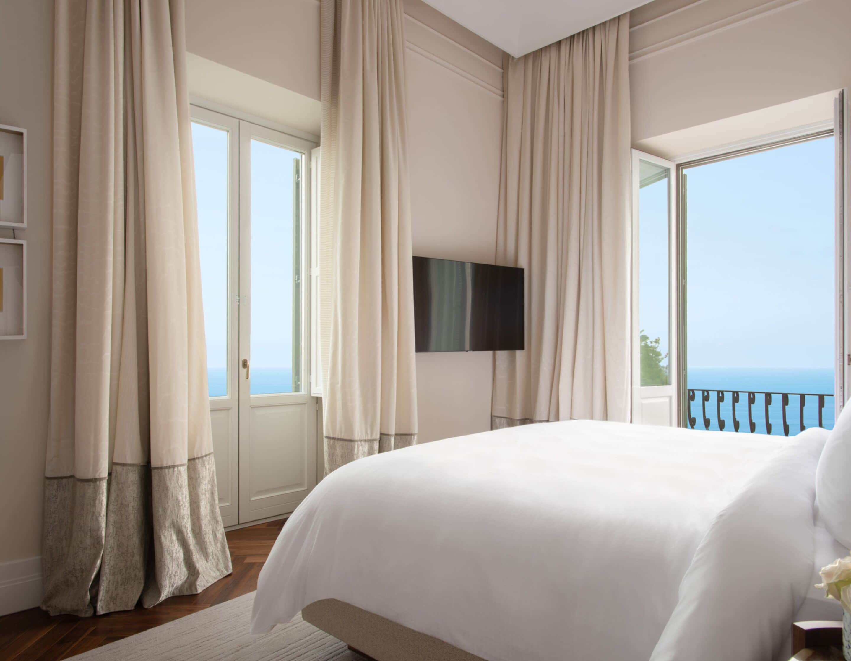 A spacious bedroom with a large bed, a television mounted on the wall, and a balcony with a view of the sea.