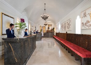 A hotel lobby with marble reception counters, a red upholstered bench, and framed artwork on the walls.