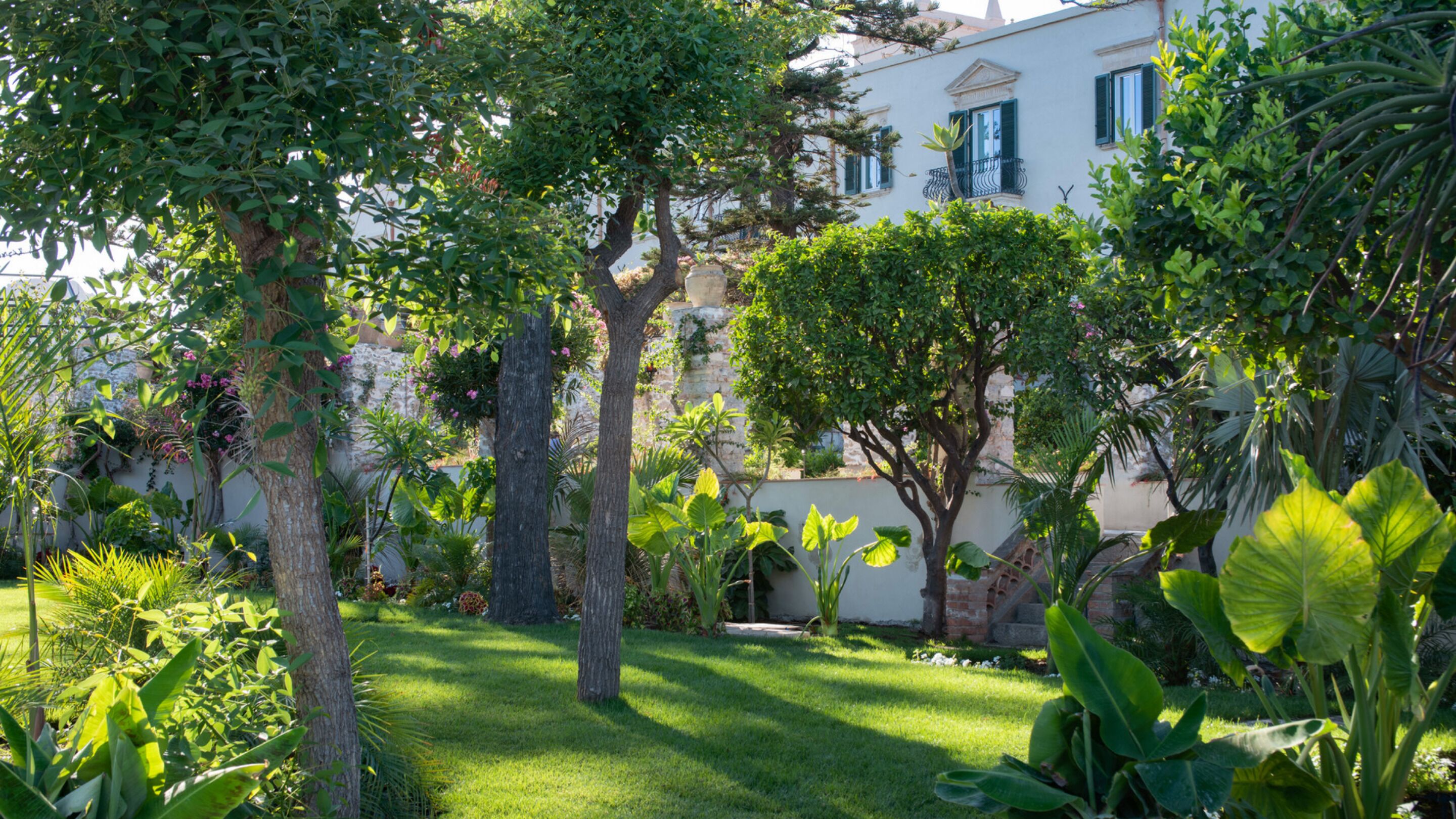 Un jardin luxuriant, avec une variété d’arbres et de plantes, une pelouse bien entretenue et un bâtiment à la façade blanche et aux volets verts.