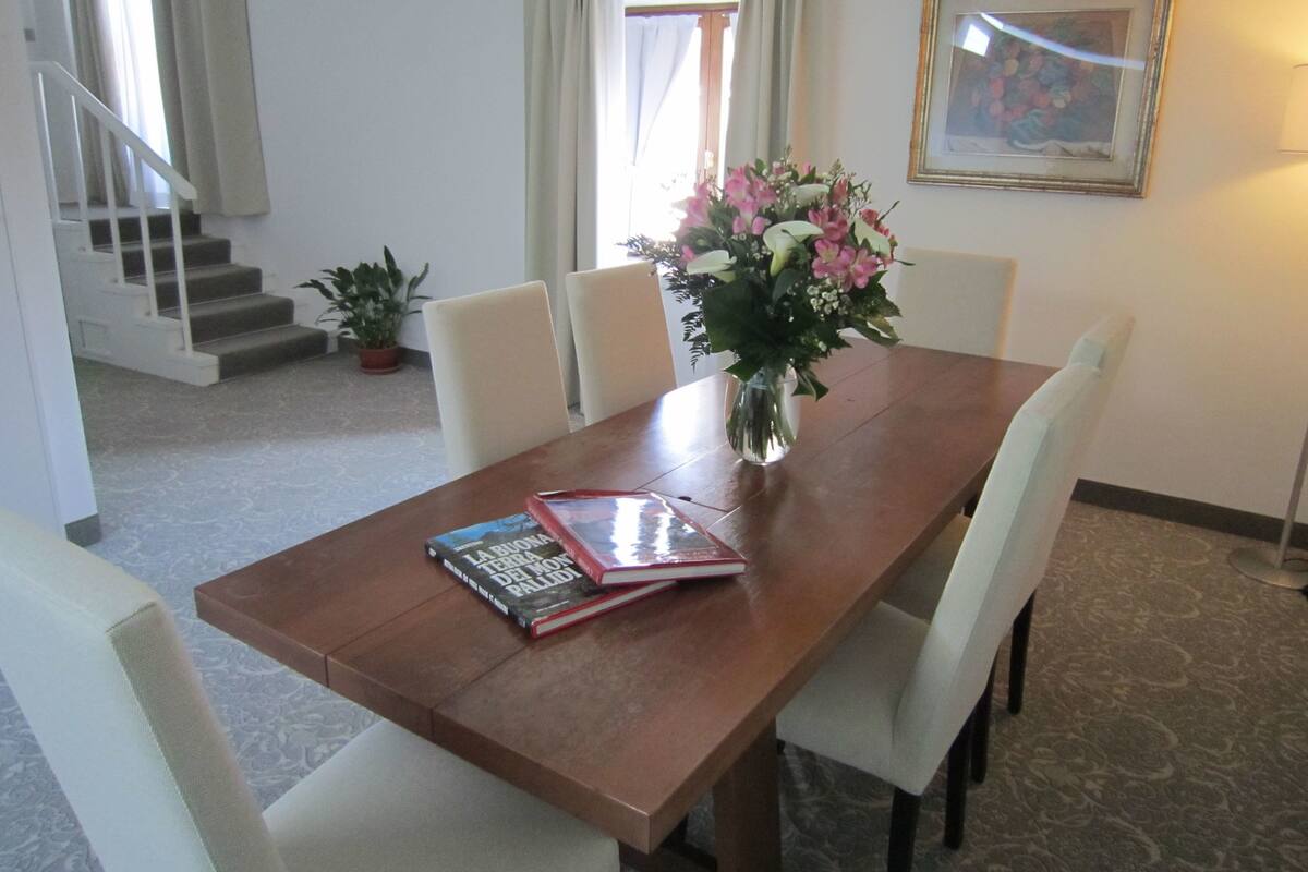 Un coin repas avec une table en bois, des chaises blanches, un vase de fleurs et un tableau encadré au mur.