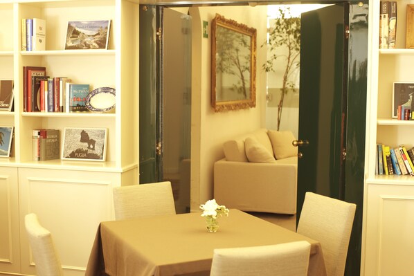 A room with a dining table, chairs, a sofa, and a bookshelf with books and framed pictures.